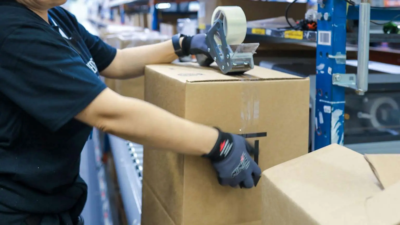 Worker taping cardboard box in warehouse with shelves and conveyor